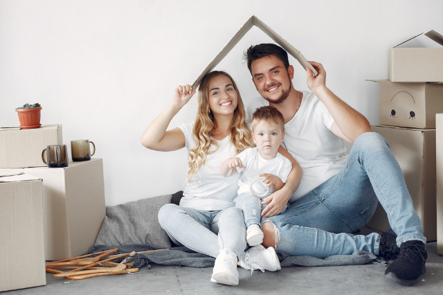 Família sorridente com uma criança pequena segurando uma casa de papelão, sentados no chão entre caixas de mudança.