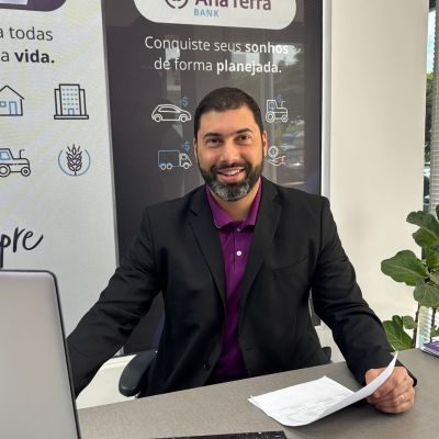 Homem sentado em uma mesa de escritório, usando terno preto e camisa roxa, com documentos e um computador à sua frente.