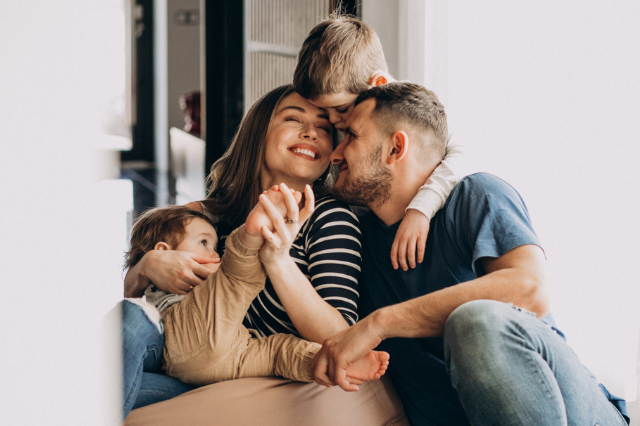 Família sorridente sentada em um sofá, com dois adultos e duas crianças, demonstrando afeto e alegria em um ambiente interno.